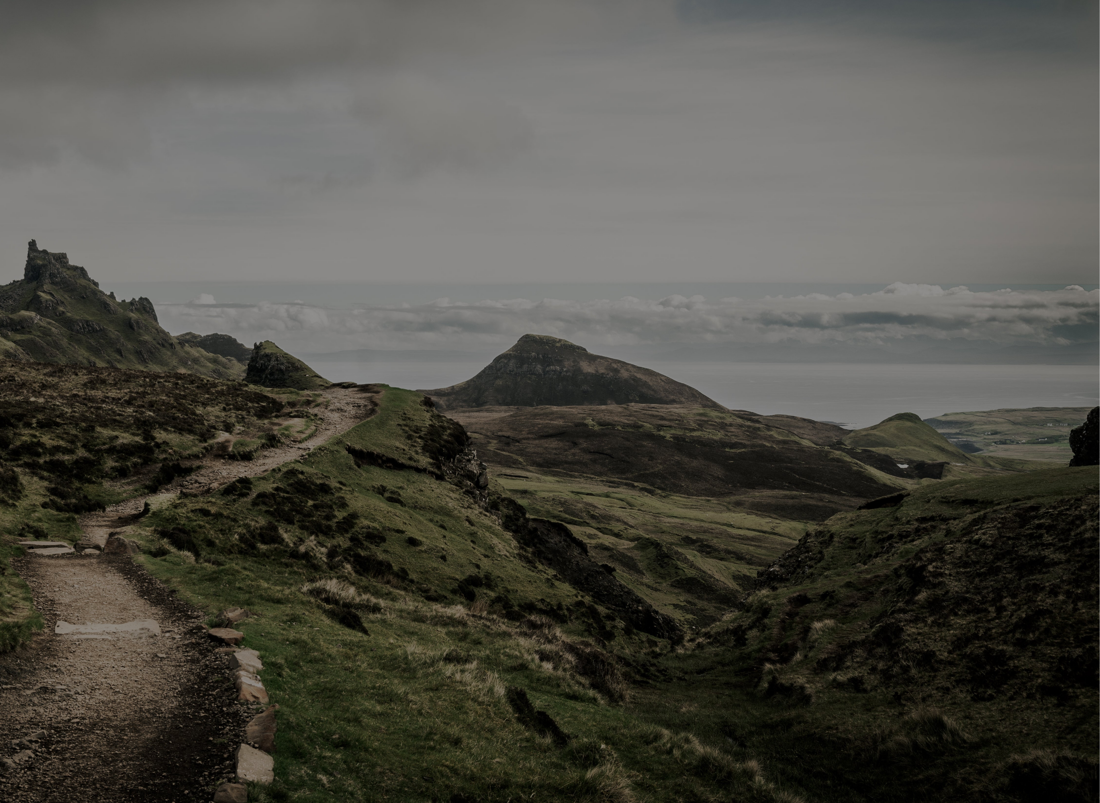 A moody, desaturated landscape photograph of a grassy mountain valley with a winding dirt path in the foreground, under a cloudy, overcast sky.